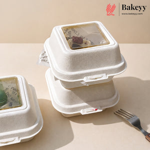 Three white biodegradable paper food containers with clear plastic lids are stacked on a beige surface. One container is open, showing a chocolate dessert inside, and another is partially visible on the left.