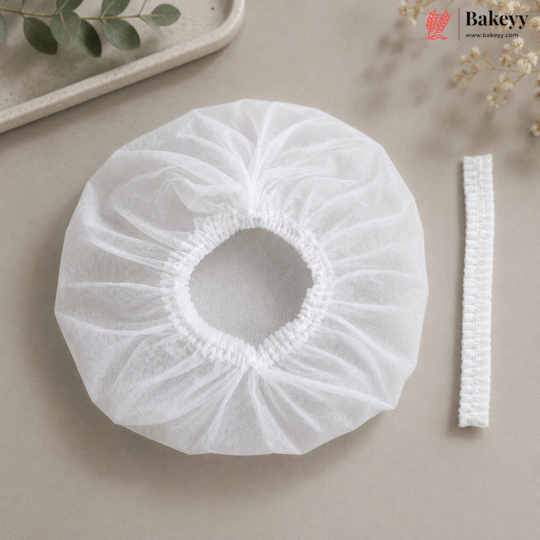A white disposable bakery dust cover with an elasticized center and a matching elastic band, displayed on a neutral beige surface with decorative greenery.