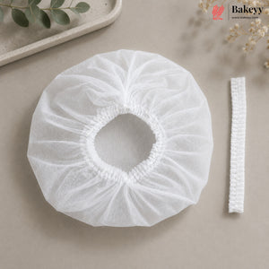 A white disposable bakery dust cover with an elasticized center and a matching elastic band, displayed on a neutral beige surface with decorative greenery.