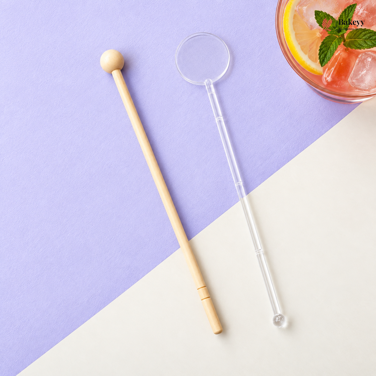 Two long cocktail straws laid on a split lavender and cream surface: one natural wood muddler with a round wooden end, and one clear plastic straw with a clear circular loop and small clear ball at the bottom. A partially visible pink drink with lemon and mint appears in the top-right corner.