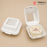Two white biodegradable square cake boxes on a beige background, with one closed and one open containing a small birthday cake. The Bakeyy logo is visible in the top right corner.