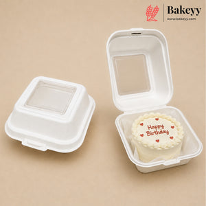Two white biodegradable square cake boxes on a beige background, with one closed and one open containing a small birthday cake. The Bakeyy logo is visible in the top right corner.