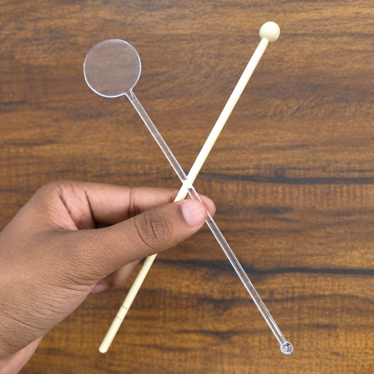 A hand holding two clear plastic straws with light beige handles, one with a circular end and one with a small rounded ball end, against a wooden background.