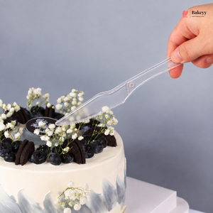A hand holding a clear plastic cake knife with serrated edges, positioned over a decorated white cake with blueberries, Oreo cookies, and white flowers against a gray background.