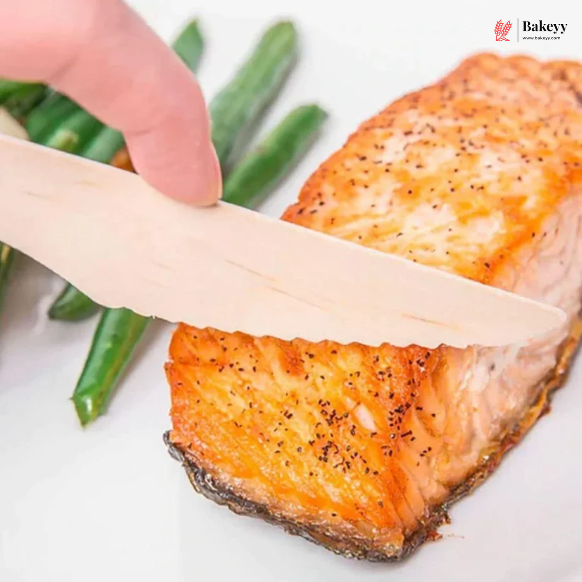 A close-up of a hand using a light beige wooden salmon fillet knife to slice a cooked salmon fillet on a white plate, with green beans in the background and a Bakeyy logo in the top right corner.
