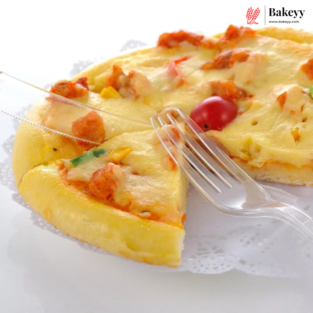 A close-up of a baked pizza with melted cheese, tomato pieces, and small vegetable bits on a white plate, with a clear plastic fork and knife and a Bakeyy logo in the top right corner.