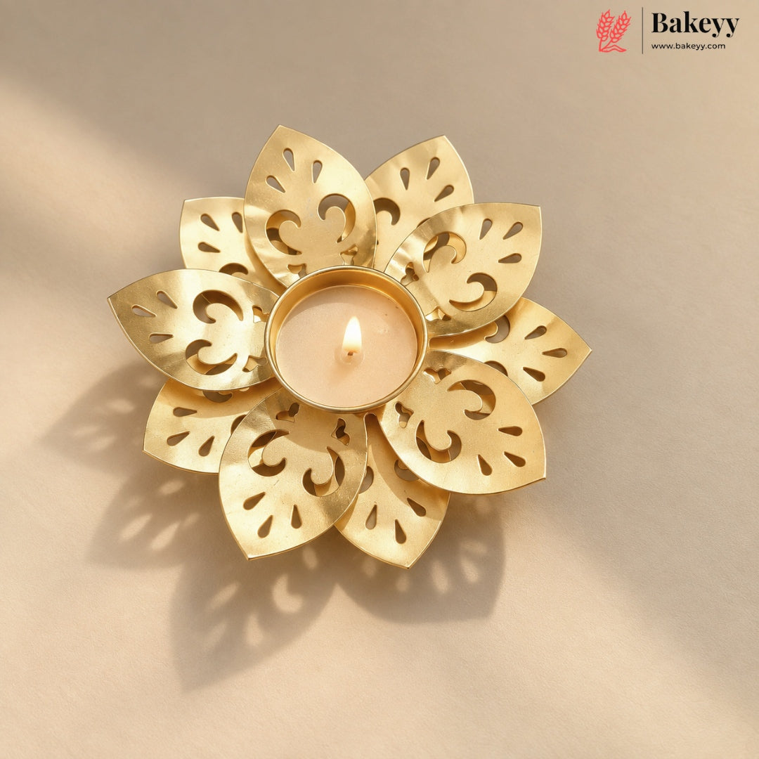 A gold-toned metal flower-shaped candle holder with cutout petal details, holding a lit tealight candle, set against a warm beige background with soft shadows. The Bakeyy logo is visible in the top right corner.