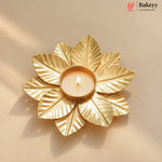 A gold-toned metal leaf-shaped candle holder with a lit tealight candle in the center, set against a warm beige background with soft shadows. The Bakeyy logo is visible in the top right corner.