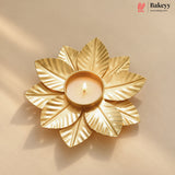 A gold-toned metal leaf-shaped candle holder with a lit tealight candle in the center, set against a warm beige background with soft shadows. The Bakeyy logo is visible in the top right corner.