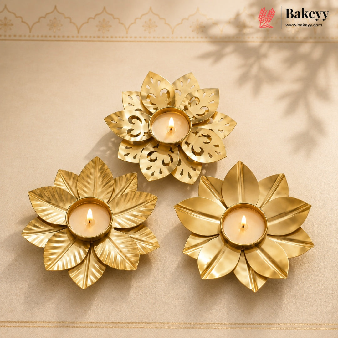 Three gold-toned flower-shaped candle holders with lit tealight candles, arranged on a beige surface with a decorative border and soft leaf shadows.