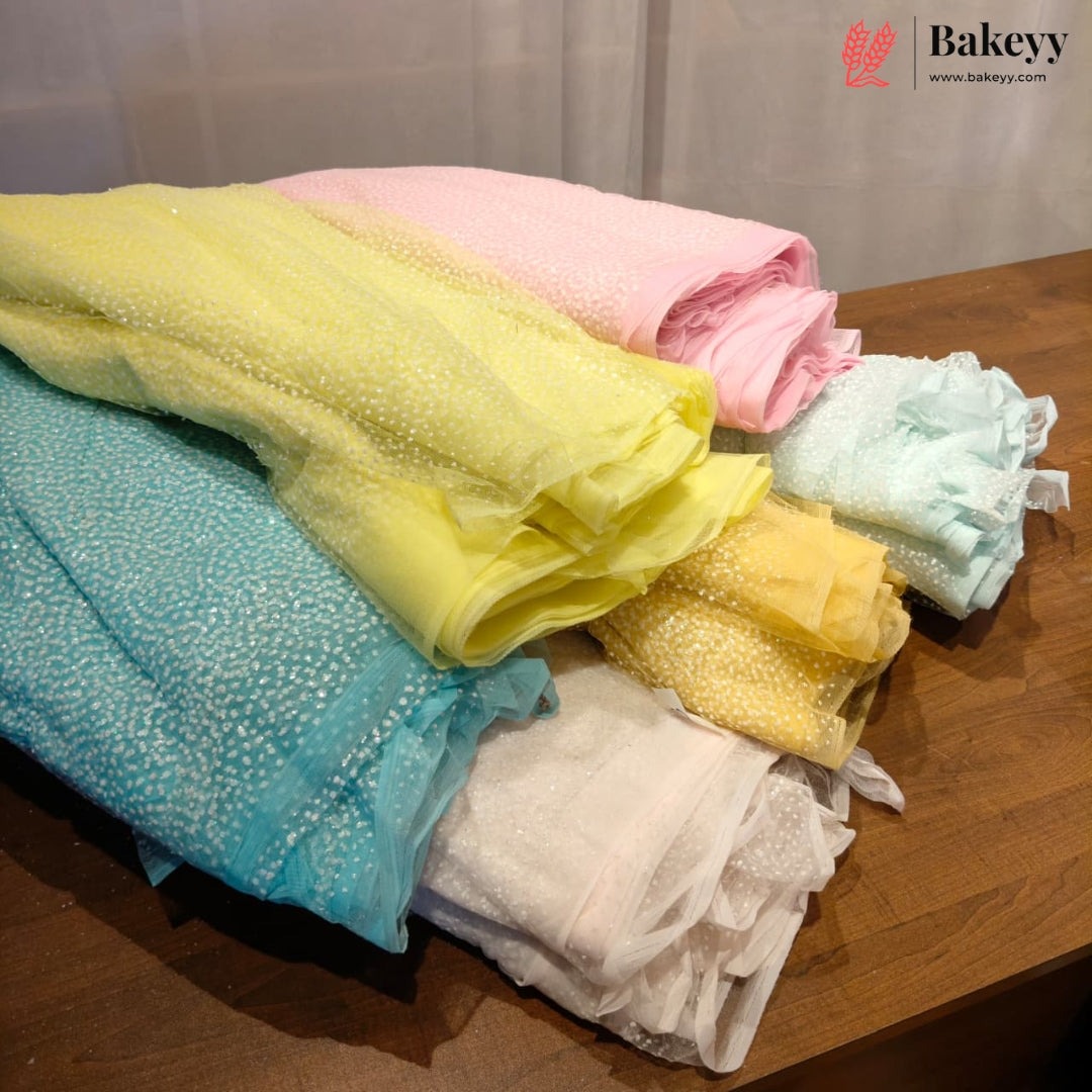 A stack of colorful ruffled lace fabric napkins in pastel shades of yellow, pink, light blue, white, and turquoise, displayed on a wooden table with a Bakeyy logo in the top right corner.