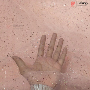 A hand placed over a sheet of pink tulle fabric with scattered glitter, showing the texture and sparkle of the material. The Bakeyy logo is visible in the top right corner.