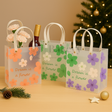 Three decorative gift bags with floral designs and text, placed on a surface with a Christmas tree in the background.
