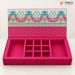 An open pink jewelry box with multiple compartments and a decorative patterned lid, photographed on a clean white background. The Bakeyy logo appears in the top right corner.