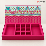 An open pink jewelry box with multiple compartments and a decorative patterned lid, photographed on a clean white background. The Bakeyy logo appears in the top right corner.