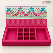 An open pink jewelry box with multiple compartments and a decorative patterned lid, photographed on a clean white background. The Bakeyy logo appears in the top right corner.