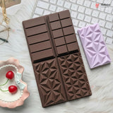 A chocolate-shaped silicone baking mold in dark brown, shown next to a lavender-colored silicone mold on a white faux fur surface with a keyboard and cherry dish as props.