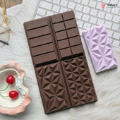A chocolate-shaped silicone baking mold in dark brown, shown next to a lavender-colored silicone mold on a white faux fur surface with a keyboard and cherry dish as props.