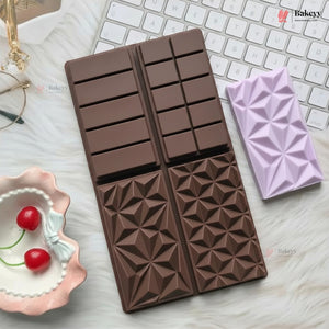A chocolate-shaped silicone baking mold in dark brown, shown next to a lavender-colored silicone mold on a white faux fur surface with a keyboard and cherry dish as props.
