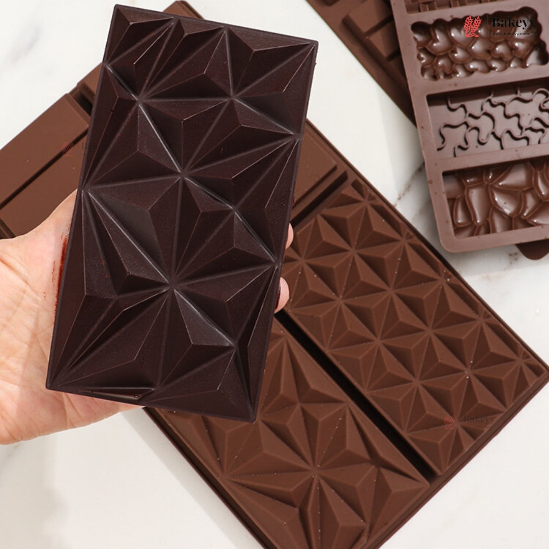 A hand holds a dark brown chocolate mold or chocolate bar with a raised geometric star pattern, with additional brown chocolate molds and a chocolate tray visible on a light marble-like surface.