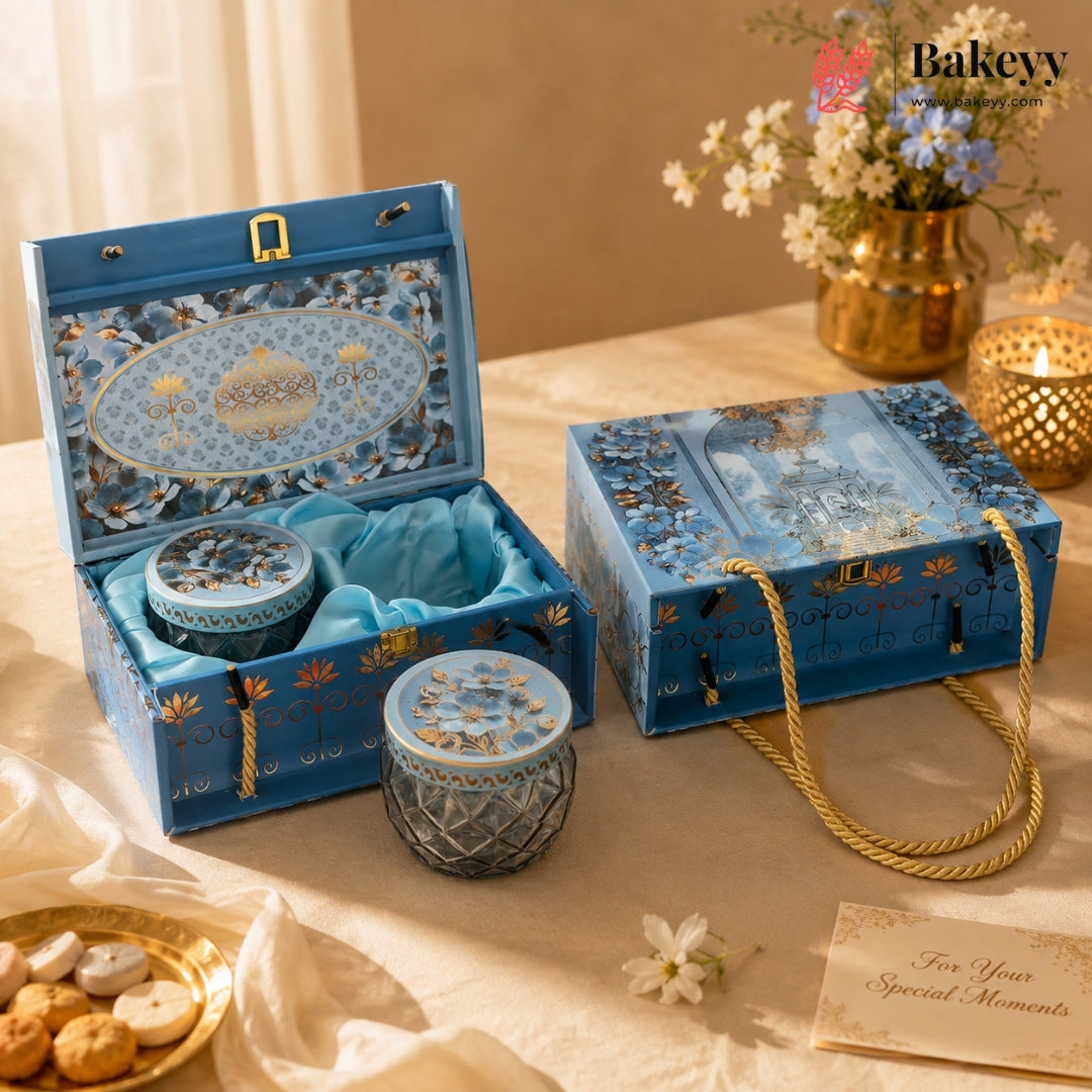 A blue decorative gift set box with floral patterns and gold accents, shown open and closed on a draped beige fabric surface. The set includes a small round tin and a textured glass jar with matching lids, styled with flowers, candles, and a small plate of cookies.