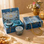A blue decorative gift set box with floral patterns and gold accents, shown open and closed on a draped beige fabric surface. The set includes a small round tin and a textured glass jar with matching lids, styled with flowers, candles, and a small plate of cookies.