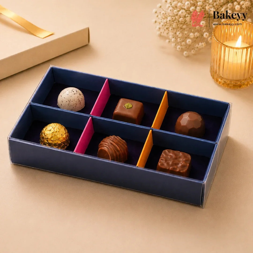 A boxed assortment of gourmet chocolates in a navy blue tray with pink and orange dividers, styled on a beige surface with a candle, dried flowers, and a gift box in the background. The Bakeey logo appears in the top right.