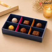 A boxed assortment of gourmet chocolates in a navy blue tray with pink and orange dividers, styled on a beige surface with a candle, dried flowers, and a gift box in the background. The Bakeey logo appears in the top right.