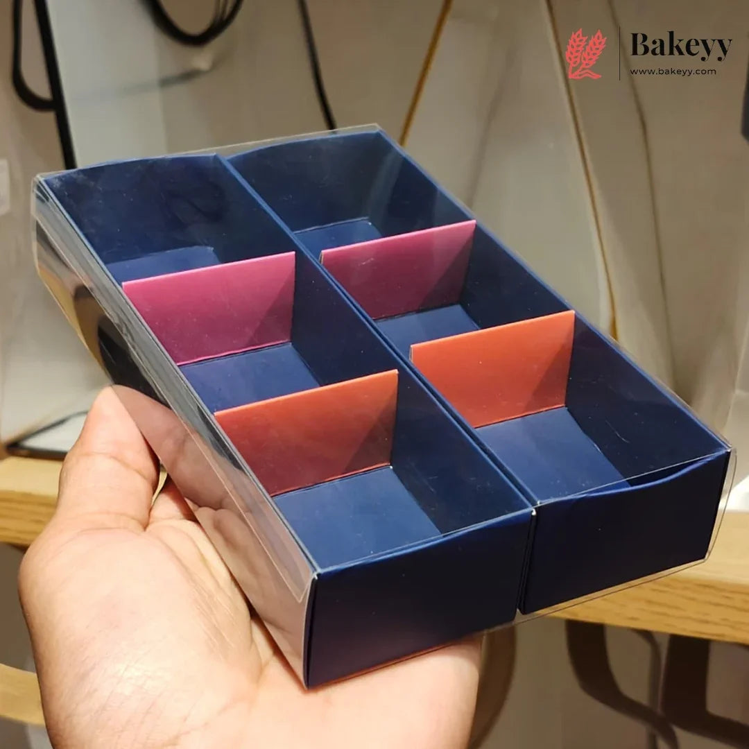 A hand holds a clear plastic divided display box with six compartments, lined with dark blue paper and accented by pink, orange, and red paper dividers. A 'Bakeyy' logo is visible in the top right corner.