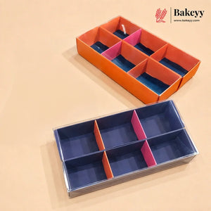 A six-compartment bakery box shown in two color configurations: one with an orange exterior and pink and blue interior dividers, and another with a navy blue exterior and pink and orange interior dividers, displayed on a beige background with the Bakeyy logo in the top right corner.
