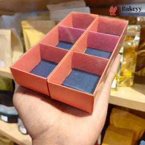 A hand holding a rectangular six-compartment cardboard tray with a peach-pink exterior and dark blue-gray interior lining, photographed in a bakery or kitchen setting with shelves and packaged goods blurred in the background. A Bakeyy watermark/logo appears in the top right.
