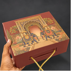 A hand holds a rectangular red gift box decorated with an ornate golden and multicolor illustration of elephants and a palace archway. The box has a gold rope handle and is photographed against a dark gray background.