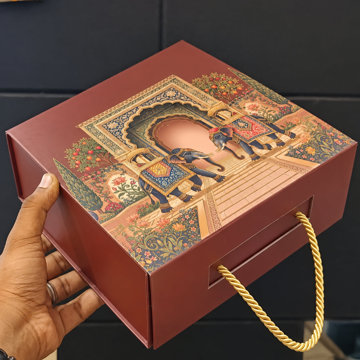 A hand holds a rectangular red gift box with a gold rope handle. The lid features an ornate, colorful illustration of elephants in a traditional architectural scene.