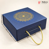 A blue rectangular bakery gift box with a gold mandala design on the lid, shown on a light beige background with dimension lines and text indicating measurements. The box has a front handle cutout with a gold rope handle and a Bakeyy logo watermark in the top right corner.