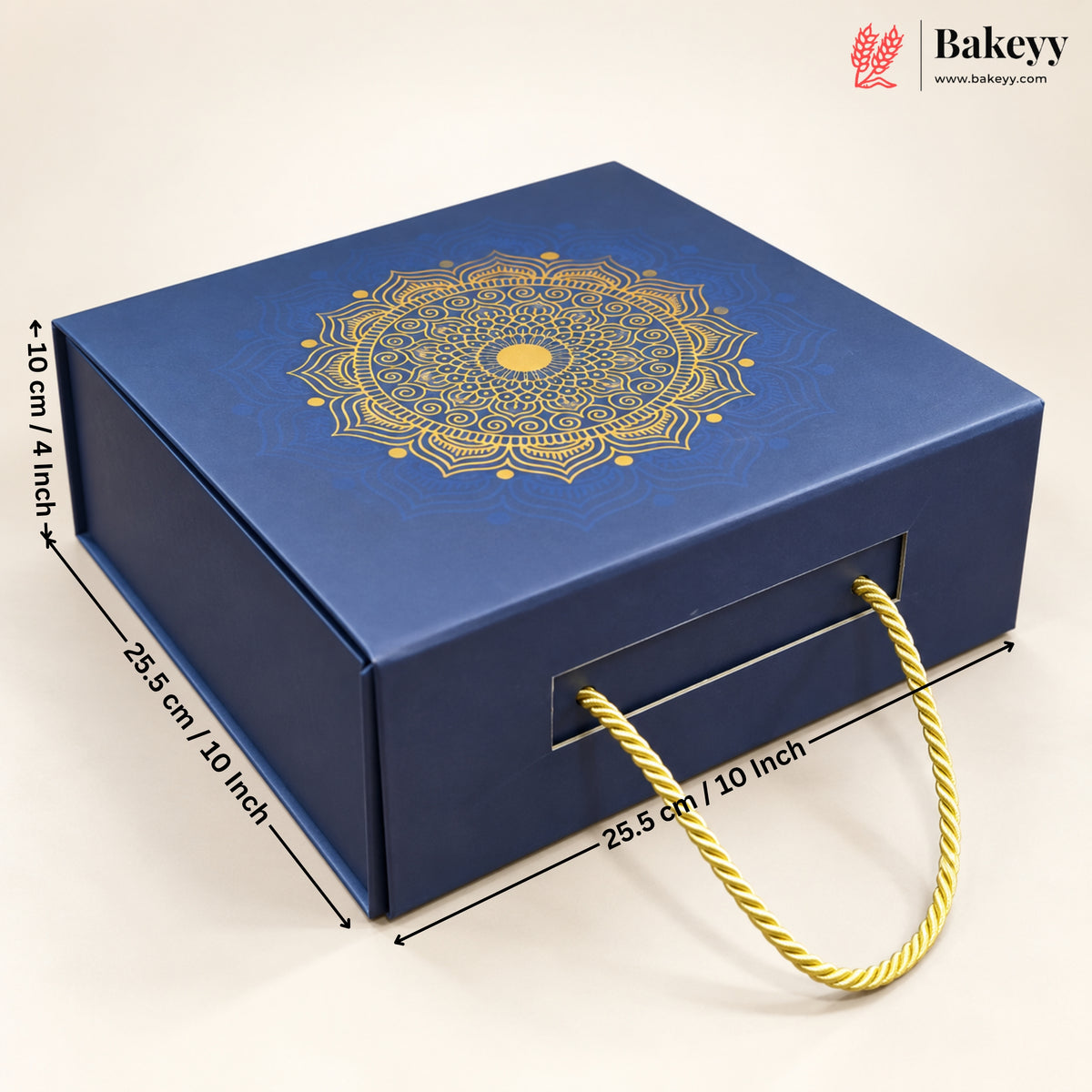 A blue rectangular bakery gift box with a gold mandala design on the lid, shown on a light beige background with dimension lines and text indicating measurements. The box has a front handle cutout with a gold rope handle and a Bakeyy logo watermark in the top right corner.