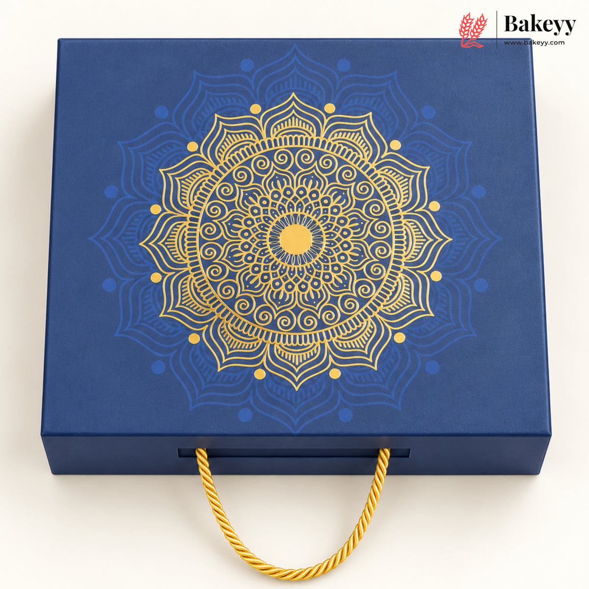 A square blue gift box with a large gold mandala design on the lid and a gold rope handle, photographed on a clean white background. A Bakeyy logo appears in the top right corner.