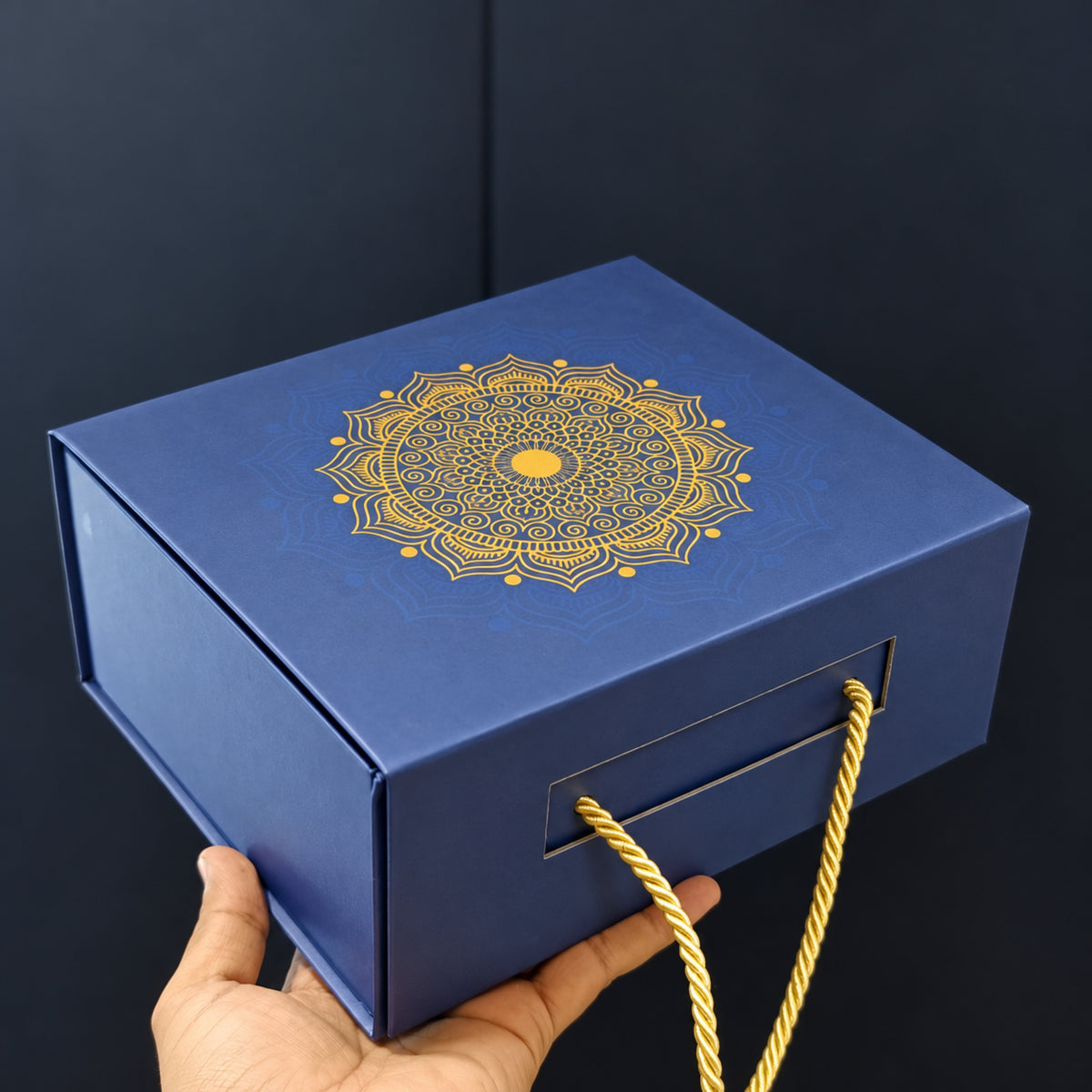 A dark blue gift box with a gold mandala design on the lid and gold rope handles, held by a hand against a dark background.