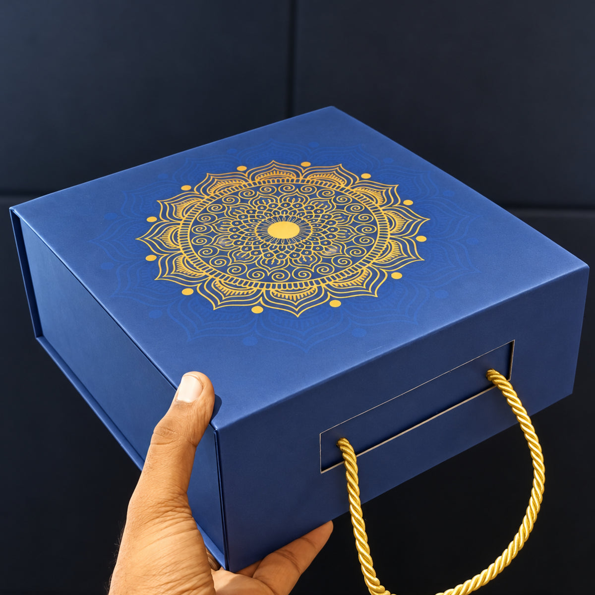 A blue gift box with a gold mandala design on the lid and a gold rope handle, held by a hand against a dark background.