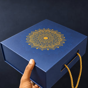 A dark blue rectangular gift box with a gold mandala design on the lid, held by a hand. The box features a gold rope handle on the side and is photographed against a dark navy background.