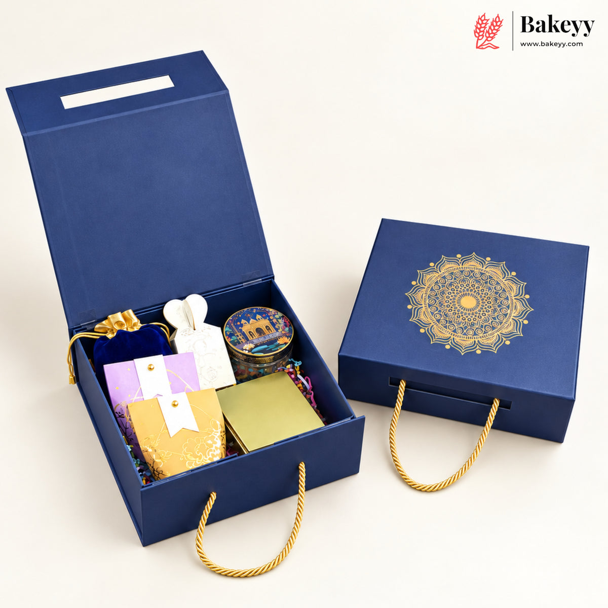 A luxury gift box set from Bakeyy, featuring an open navy blue gift box filled with assorted packaged sweets and a tin, alongside a closed matching navy blue box with a gold mandala design and gold rope handles, on a clean off-white background.