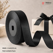 A roll of black satin ribbon unspooled across a neutral beige surface, with a gift box tied in black ribbon and decorative dried stems in the background. Text in the lower left reads 'Width: 1 Inch/2.