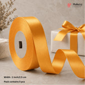 A roll of wide satin ribbon in a warm golden yellow color, unspooled across a light stone surface next to a wrapped gift box with a matching bow. Text at the bottom reads 'Width: 1 Inch/2.