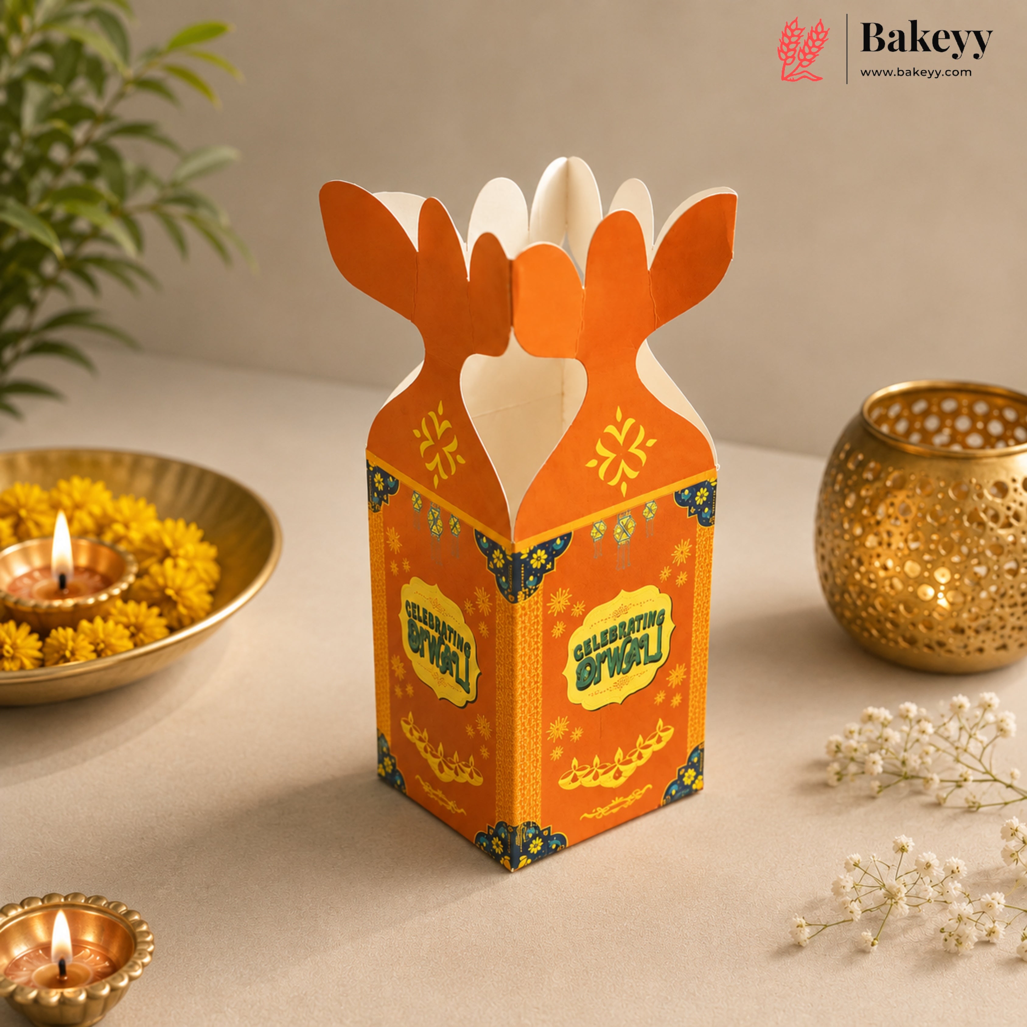 An orange and white cardboard gift box with butterfly-shaped handles and Diwali-themed decorative prints, styled on a beige surface with lit diya lamps, yellow flowers, and a gold decorative pot in the background. The Bakeyy brand logo is visible in the top right corner.