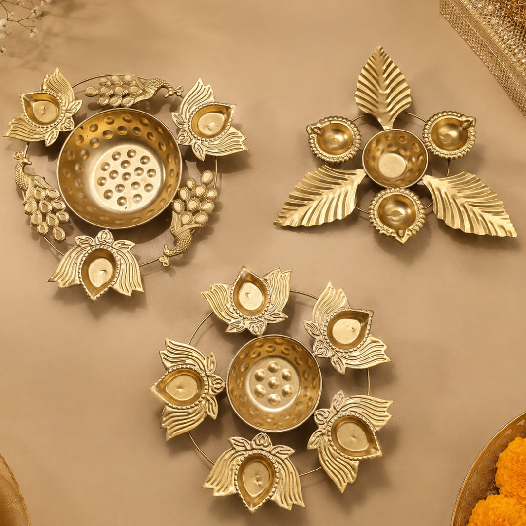 Three gold-toned brass Diwali oil lamps arranged on a beige surface, featuring decorative leaf, floral, and peacock motifs around central candle holders.