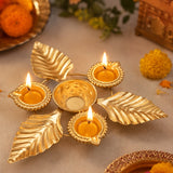 A gold-toned leaf-shaped diya holder with five candle holders, including three lit candles, arranged on a neutral surface with decorative marigold flowers and traditional ornaments in the background.
