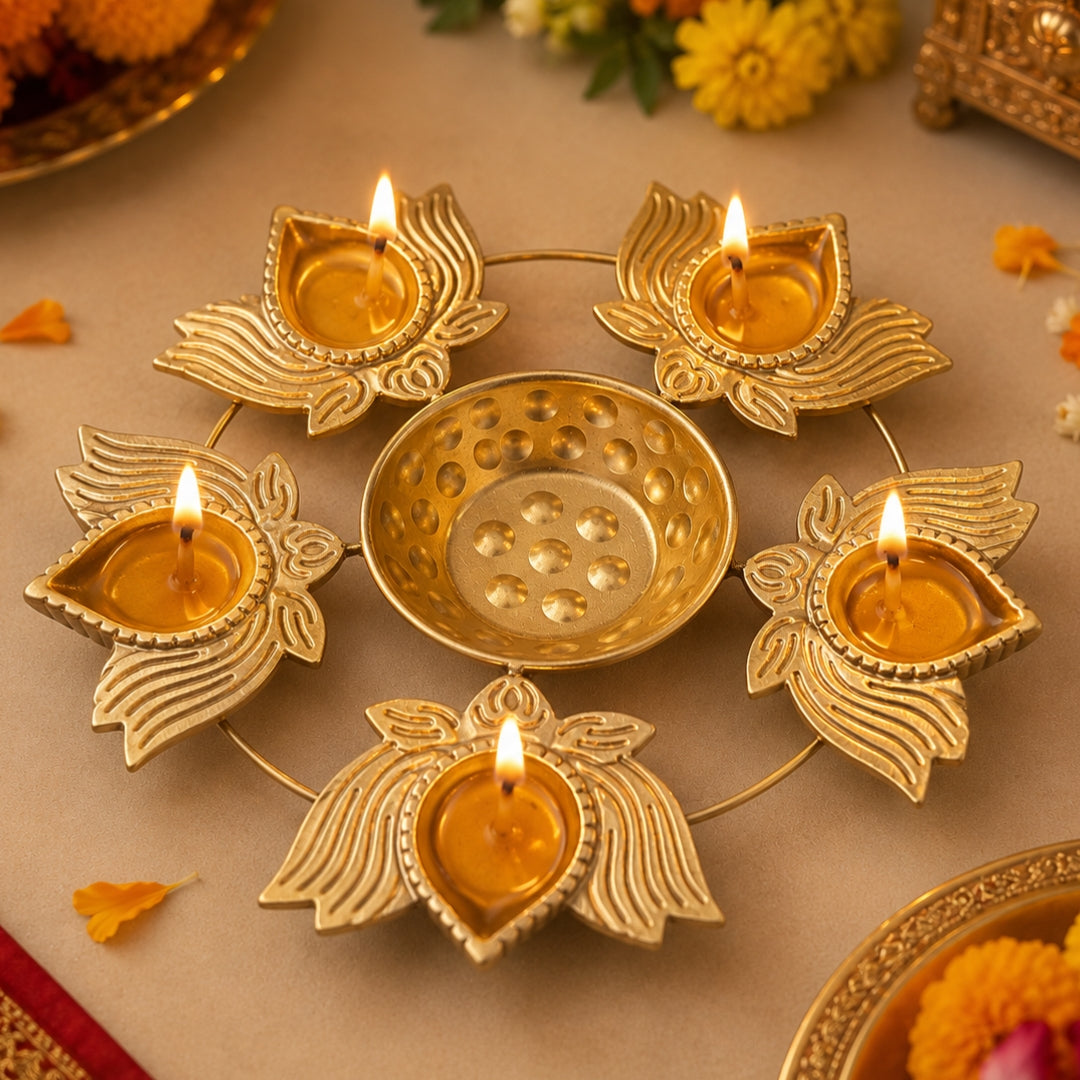 A brass diya light holder with five lotus-shaped candle holders and a central bowl, all lit with small flames, arranged on a beige surface with decorative flowers and petals.