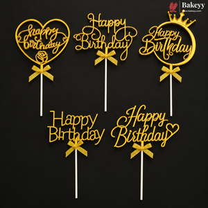 Five gold acrylic 'Happy Birthday' cake toppers with different designs including a heart, ring, and crown, each with a ribbon bow and white stick, displayed on a black background. A Bakeey brand logo is visible in the top right corner.