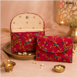 A pair of red embroidered fabric clutch bags displayed on a decorative tray with warm-toned props including a lit diya, flowers, and a brass vessel. The Bakeyy brand logo is visible in the top right corner.