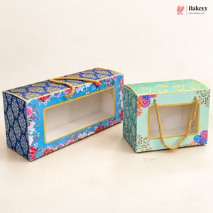 Two decorative rectangular bakery gift boxes with clear front windows and gold rope handles, shown on a plain light beige background. One box is blue with floral and ornamental patterns, and the other is mint green with similar floral detailing.