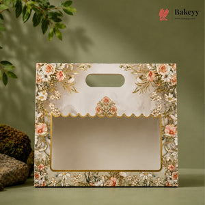 A decorative bakery gift bag with a floral print border, gold trim, and a scalloped window frame, shown on a muted green background with a leafy plant and rocks as props.